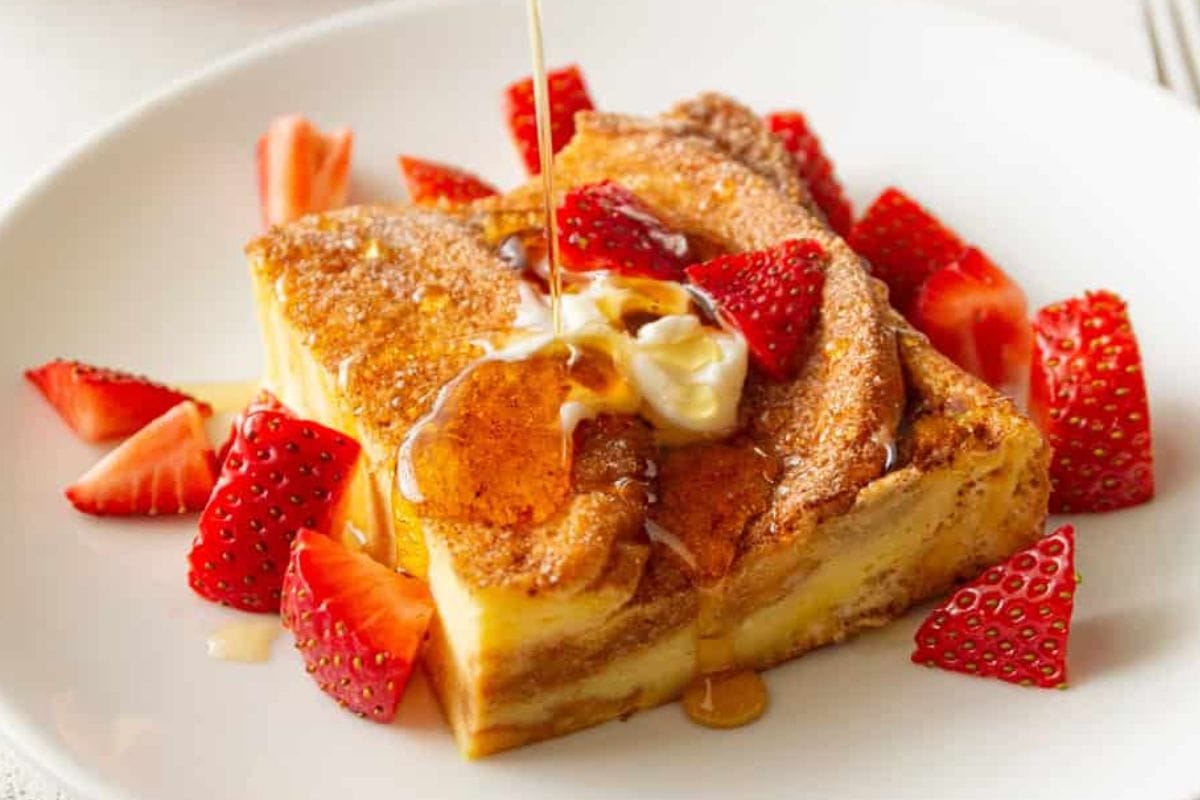 A slice of French toast topped with syrup, whipped cream, and fresh strawberries on a white plate.