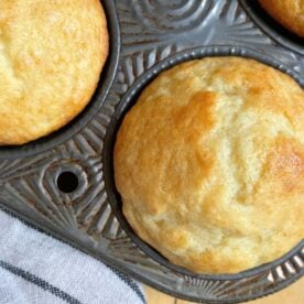 Close-up of golden-brown buttermilk muffins in a vintage muffin tin, with a striped linen cloth in the background.
