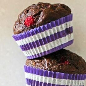Two chocolate raspberry muffins topped with a raspberry, displayed in purple and white striped cupcake liners.