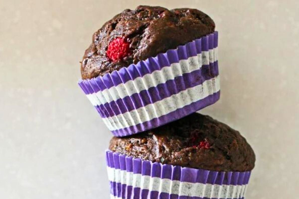 Two chocolate raspberry muffins topped with a raspberry, displayed in purple and white striped cupcake liners.