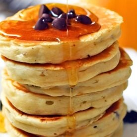 A stack of fluffy pancakes drizzled with syrup and topped with chocolate chips, on a white plate.