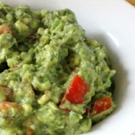 A close-up of a bowl filled with creamy guacamole featuring diced tomatoes and green herbs, served alongside tortilla chips.