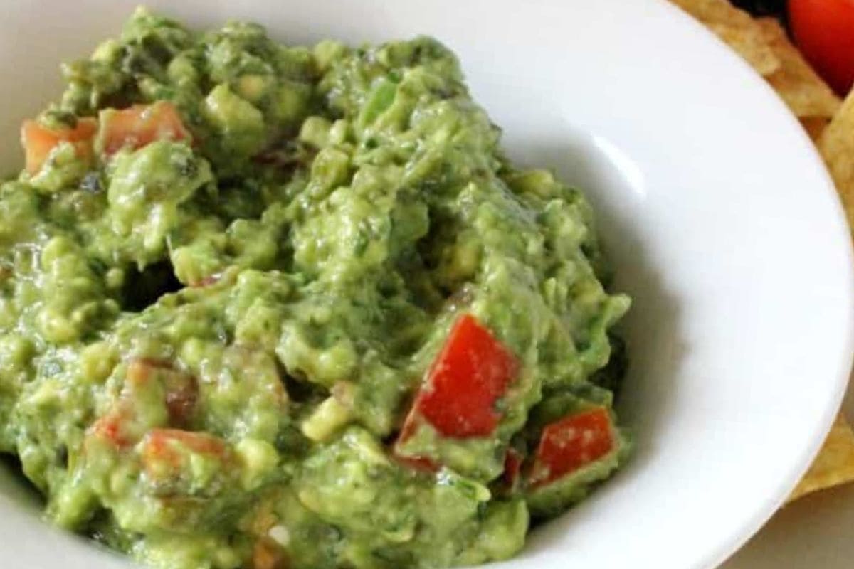 A close-up of a bowl filled with creamy guacamole featuring diced tomatoes and green herbs, served alongside tortilla chips.