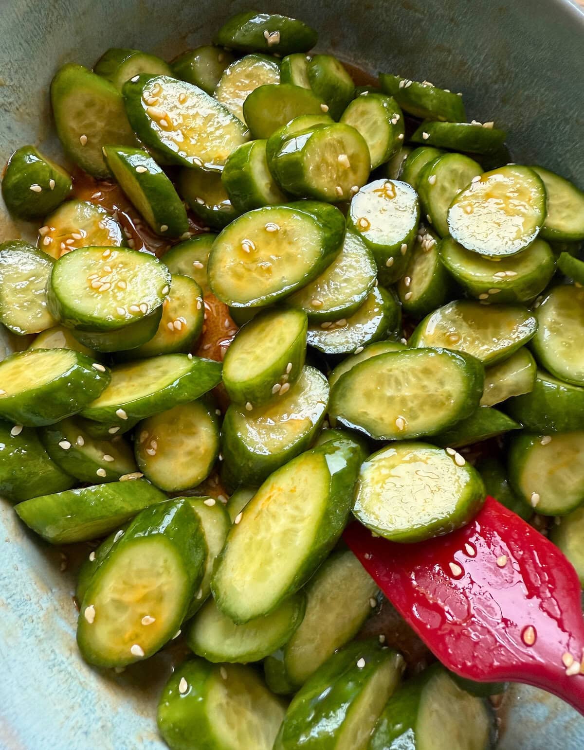 Sliced cucumbers in a bowl with sesame seeds and a red spatula, coated in a tangy sesame dressing.
