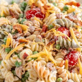 Close-up of colorful pasta salad featuring bacon, peas, cherry tomatoes, and shredded cheese in a creamy dressing.