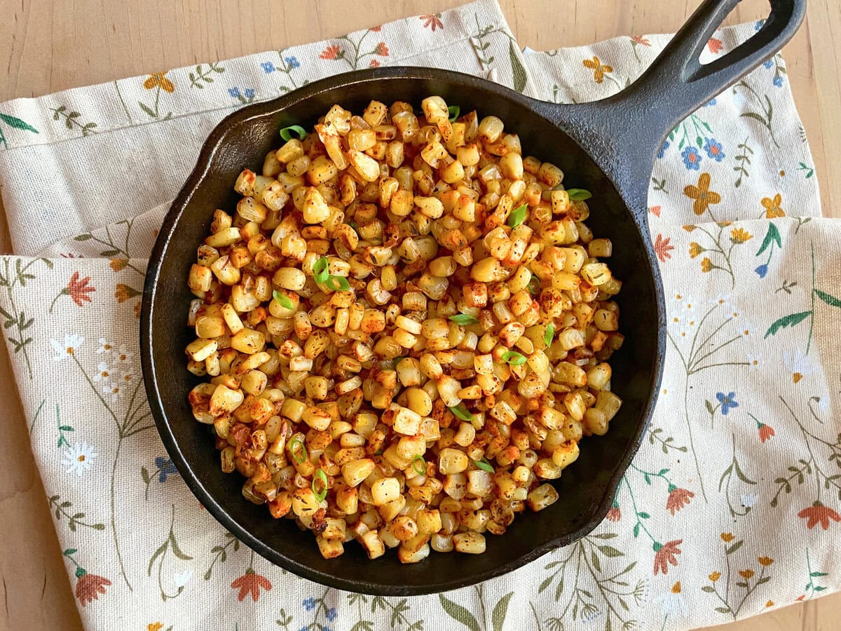 A cast iron skillet filled with blackened corn, seasoned and garnished with green onions, resting on a floral-patterned cloth.