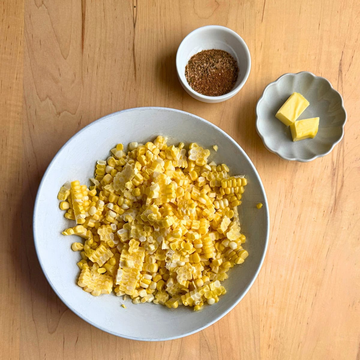A bowl of freshly cut corn kernels accompanied by a small dish of seasoning and a portion of butter on a wooden surface.