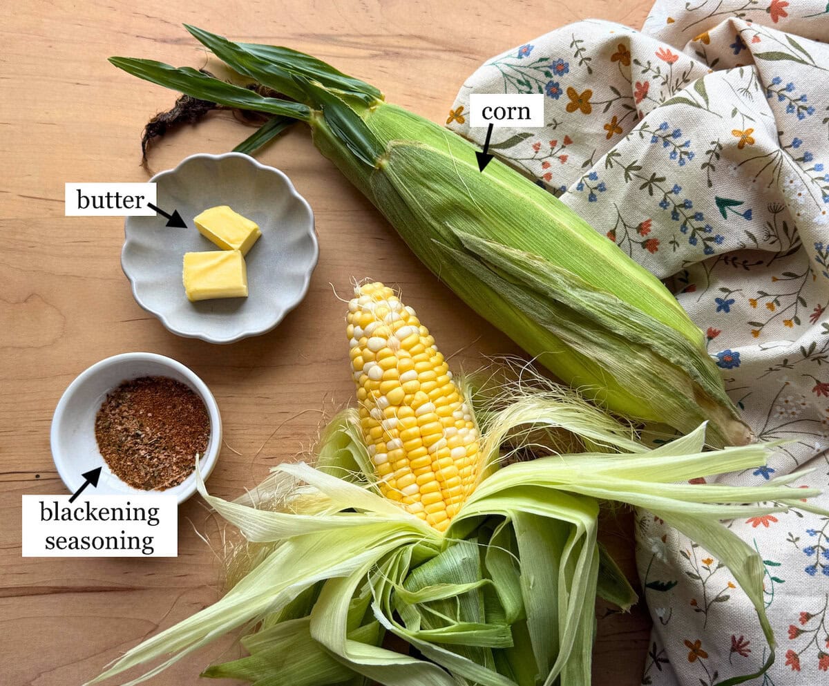 An arrangement of corn, butter, and blackening seasoning on a wooden surface, with a floral-patterned cloth in the background.