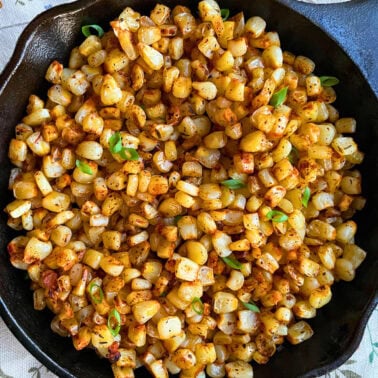 A close-up of blackened, sautéed corn kernels garnished with fresh green onions, served in a cast iron skillet.
