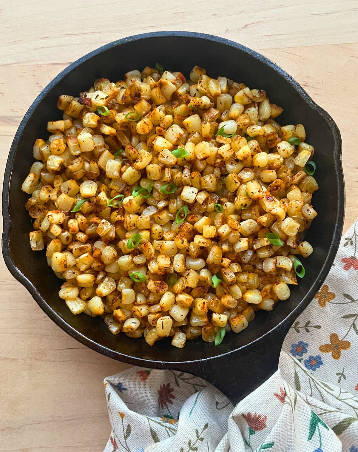 A skillet filled with golden blackened corn kernels topped with chopped green onions, resting on a floral napkin.