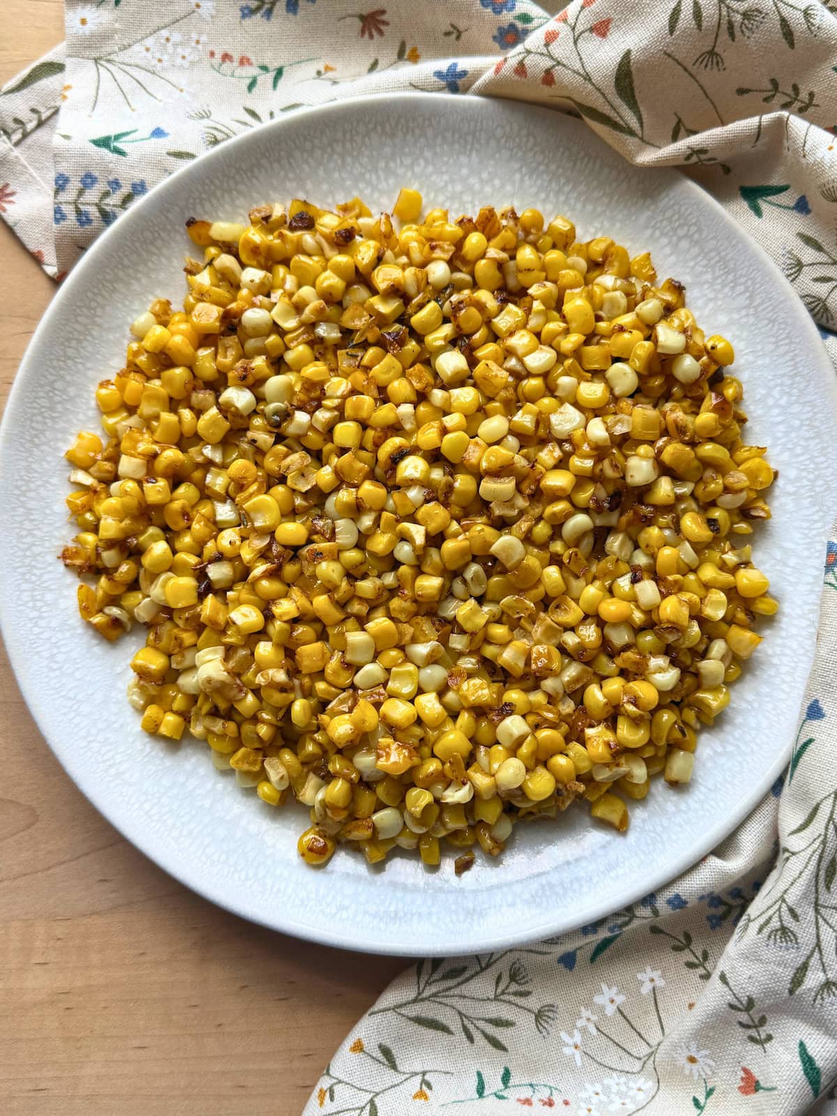 A plate of blackened, golden corn kernels served on a textured white plate, with a floral-patterned cloth in the background.