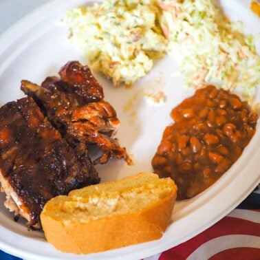 A hearty plate of BBQ ribs, coleslaw, baked beans, and a slice of cornbread, set against a backdrop of a patriotic flag.
