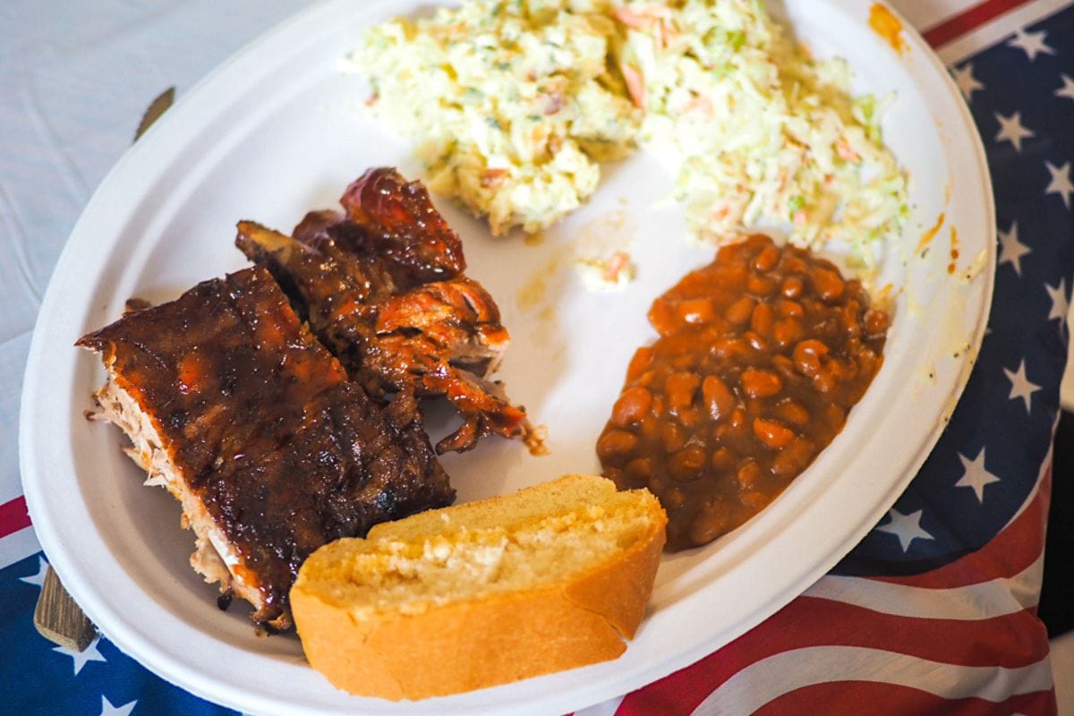 A hearty plate of BBQ ribs, coleslaw, baked beans, and a slice of cornbread, set against a backdrop of a patriotic flag.
