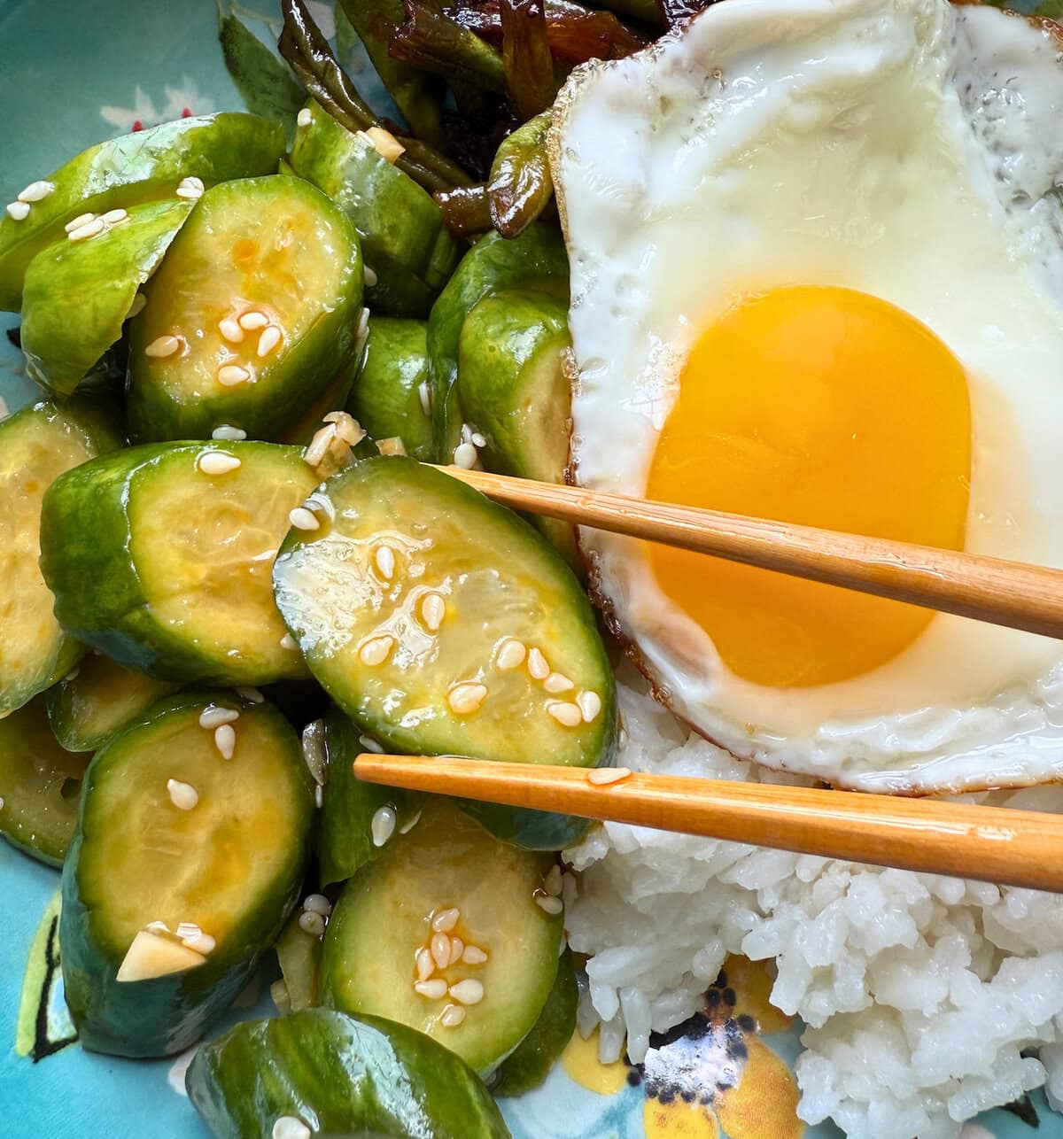 A close-up of spicy Asian cucumber salad, accompanied by a fried egg and rice, with chopsticks resting on the dish.