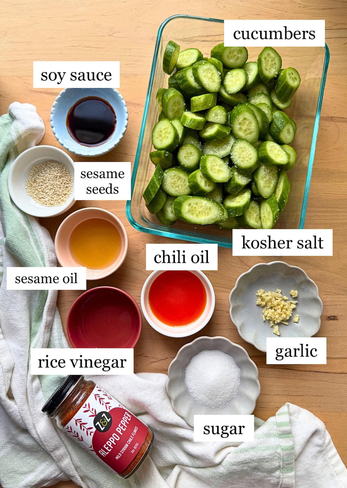 Sliced cucumbers accompanied by various condiments and seasonings including soy sauce, chili oil, sesame oil, rice vinegar, garlic, sugar, and sesame seeds.