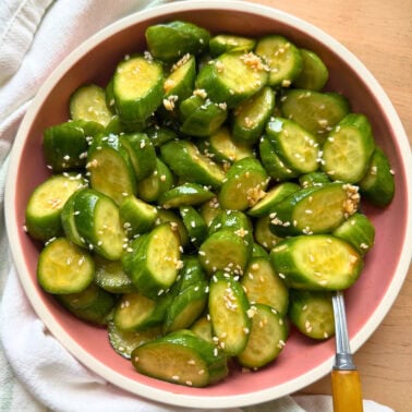 Freshly sliced cucumbers topped with sesame seeds in a pink bowl, set against a neutral background.