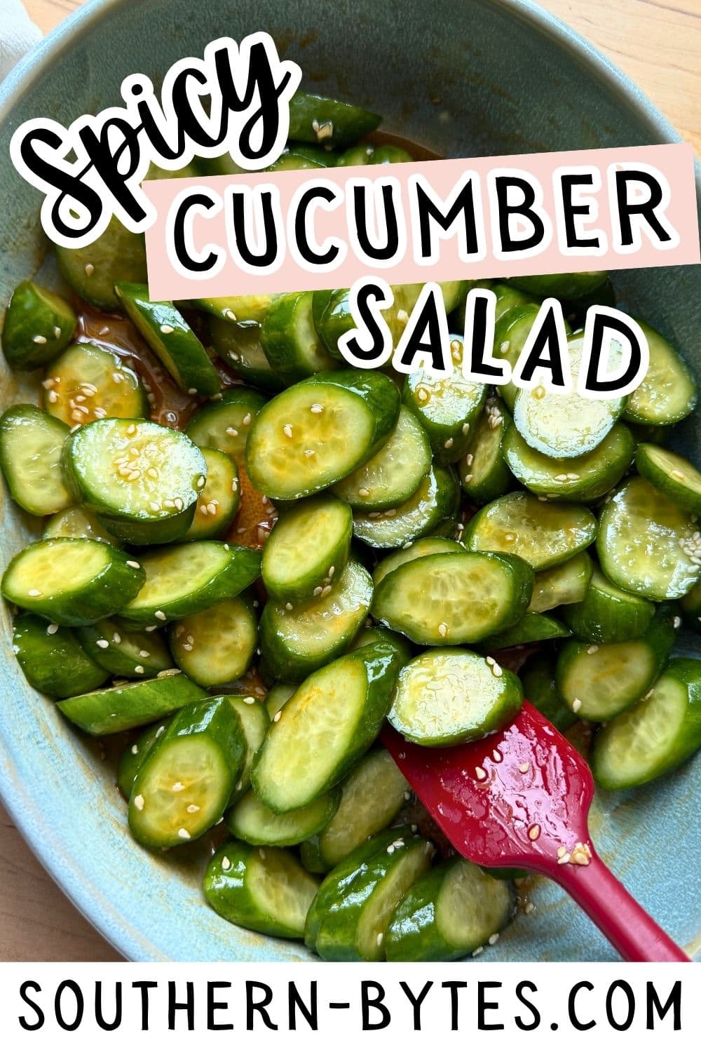 A colorful bowl filled with sliced cucumbers in a spicy dressing, topped with sesame seeds and a red spatula for serving.
