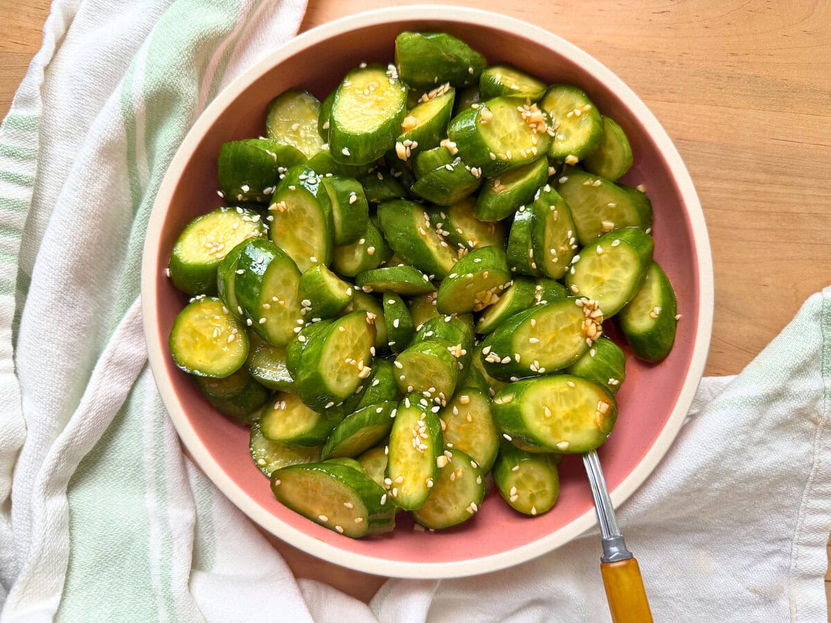 A pink bowl of spicy Asian cucumber salad topped with sesame seeds, served on a light background with a textured napkin.