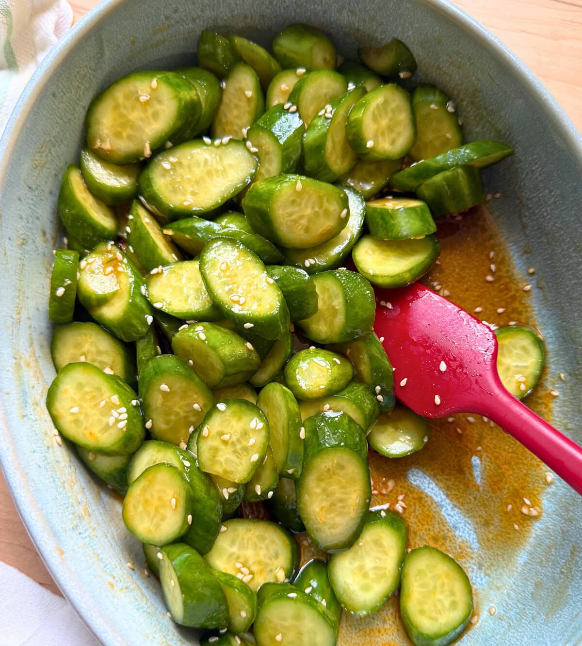 A vibrant bowl of spicy cucumber salad with sesame dressing, featuring a red spatula resting in the bowl.