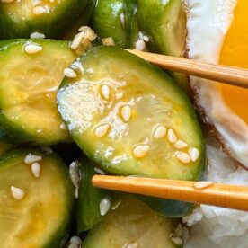 Close-up of sliced cucumbers in a sesame dressing with a fried egg and chopsticks on white rice.
