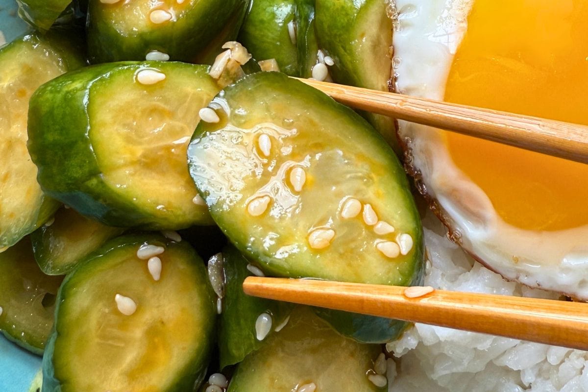Close-up of sliced cucumbers in a sesame dressing with a fried egg and chopsticks on white rice.