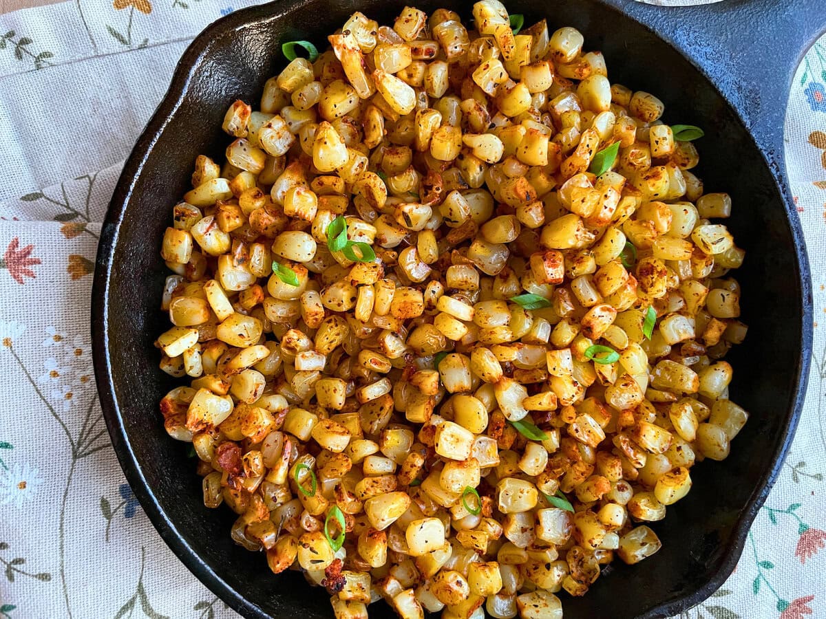 A close-up of golden skillet-roasted corn garnished with green onions, presented in a cast iron skillet on a floral patterned tablecloth.