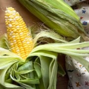 An ear of fresh sweet corn surrounded by green husks, resting on a wooden surface alongside a floral-patterned cloth.