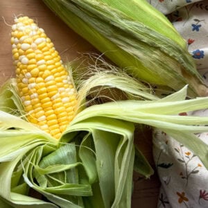 An ear of fresh sweet corn surrounded by green husks, resting on a wooden surface alongside a floral-patterned cloth.