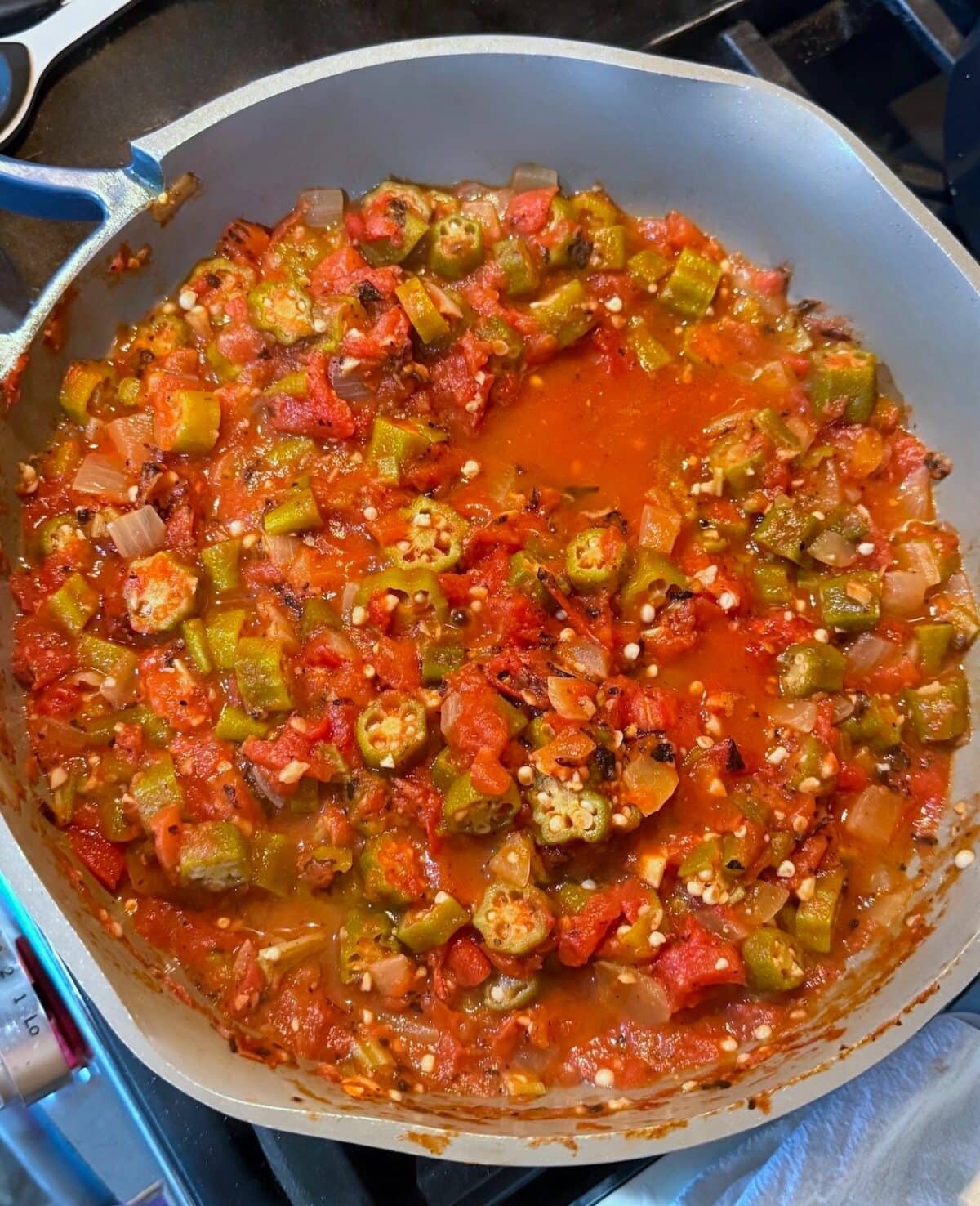 A close-up view of a cooked vegetable medley featuring okra, diced tomatoes, and onions in a vibrant red sauce, simmering in a large pot on the stovetop.