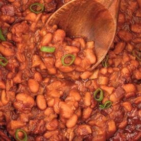 Close-up of rich baked beans with green onion garnish, served in a pot with a wooden spoon.