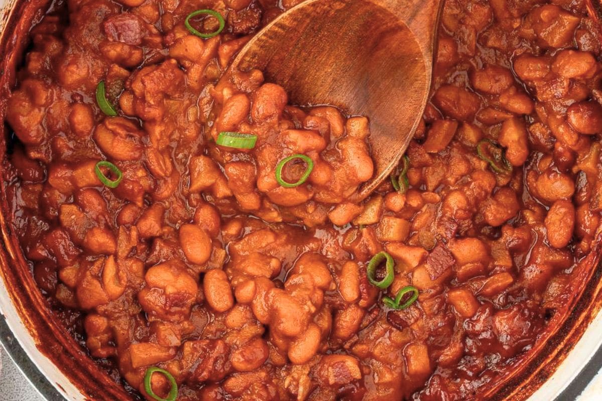 Close-up of rich baked beans with green onion garnish, served in a pot with a wooden spoon.