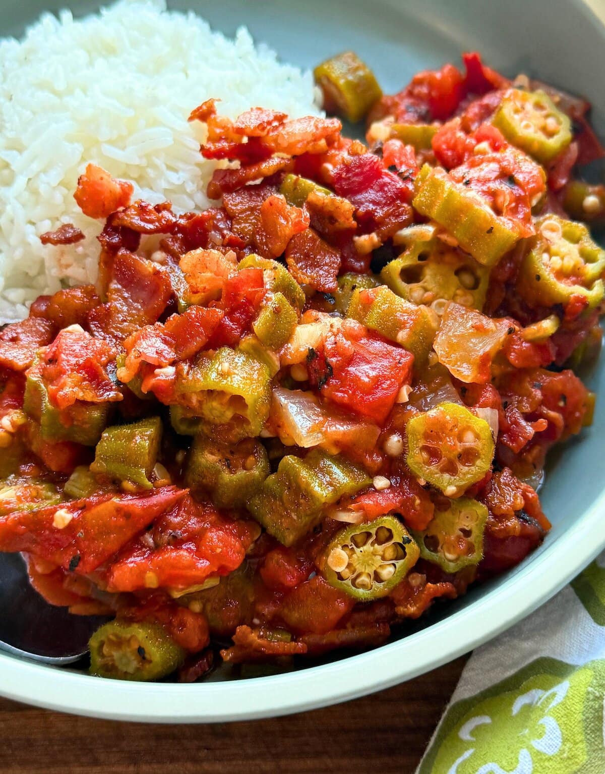 A plate featuring hearty stewed okra and tomatoes with bacon paired with a generous portion of fluffy white rice, highlighting vibrant red tomatoes and green okra pieces.