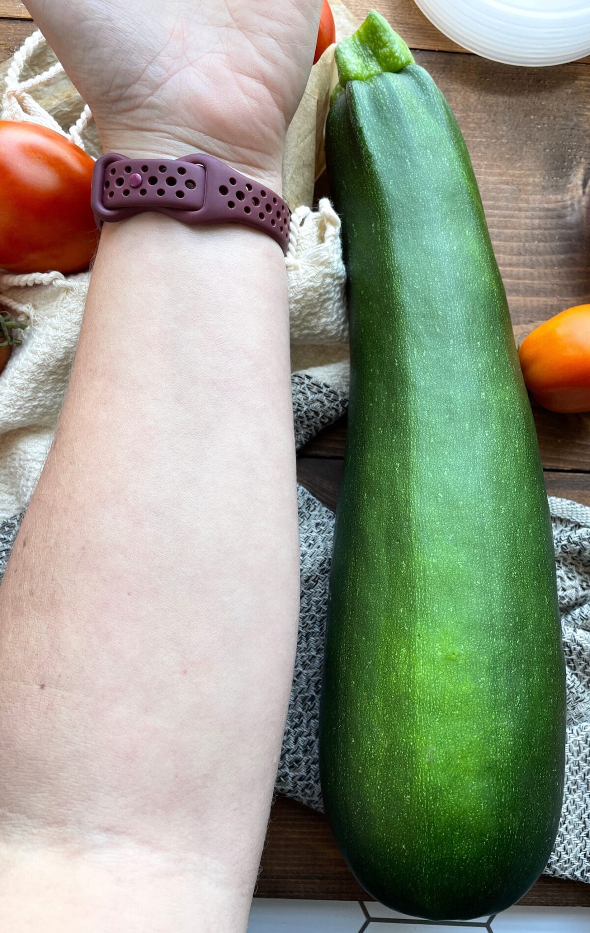 A close-up view of a large zucchini next to a hand, showcasing the scale with tomatoes and kitchen linens in the background.