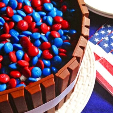 A delicious chocolate cake topped with red and blue chocolate candies, surrounded by Kit Kat bars, alongside American flag napkins.