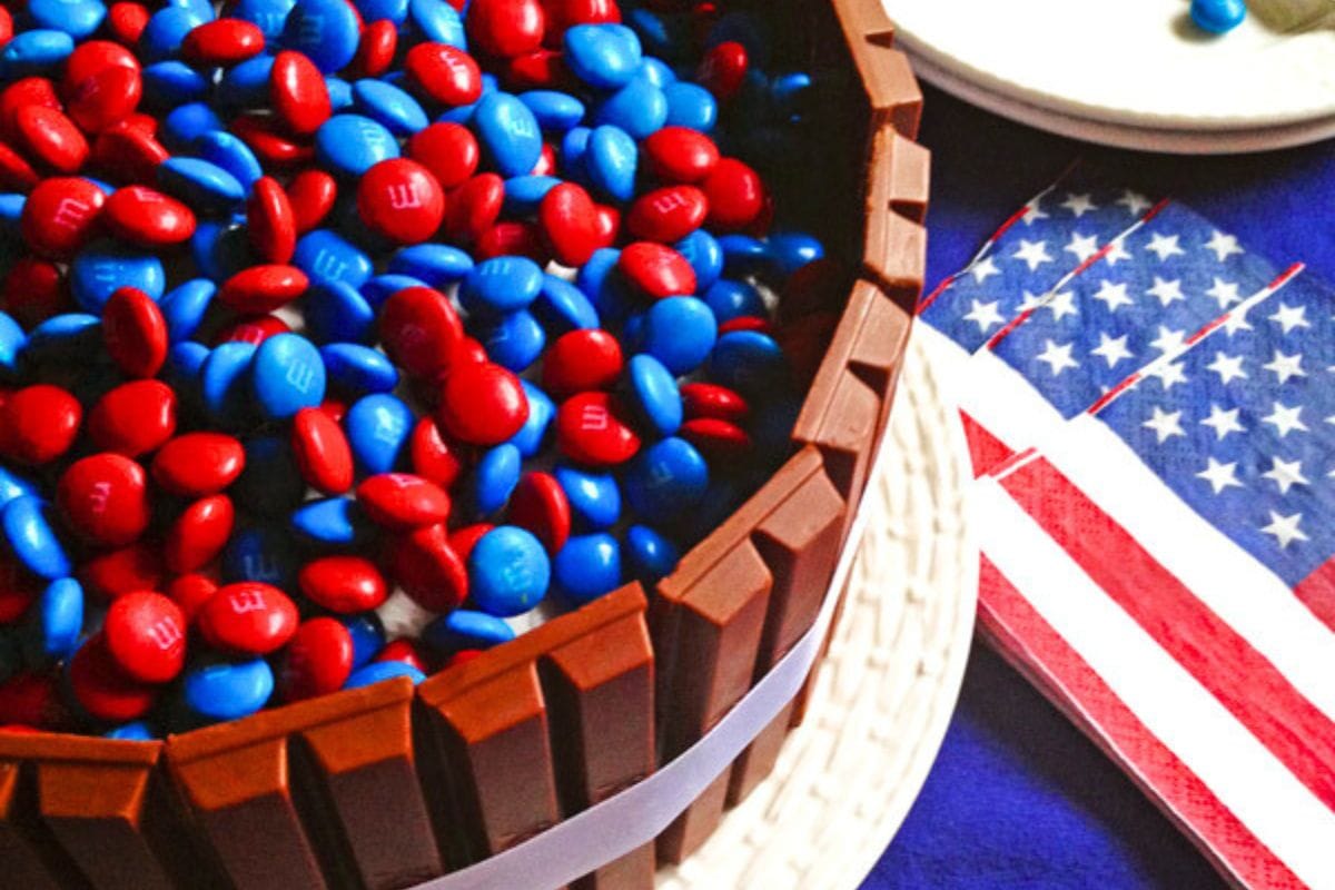 A delicious chocolate cake topped with red and blue chocolate candies, surrounded by Kit Kat bars, alongside American flag napkins.