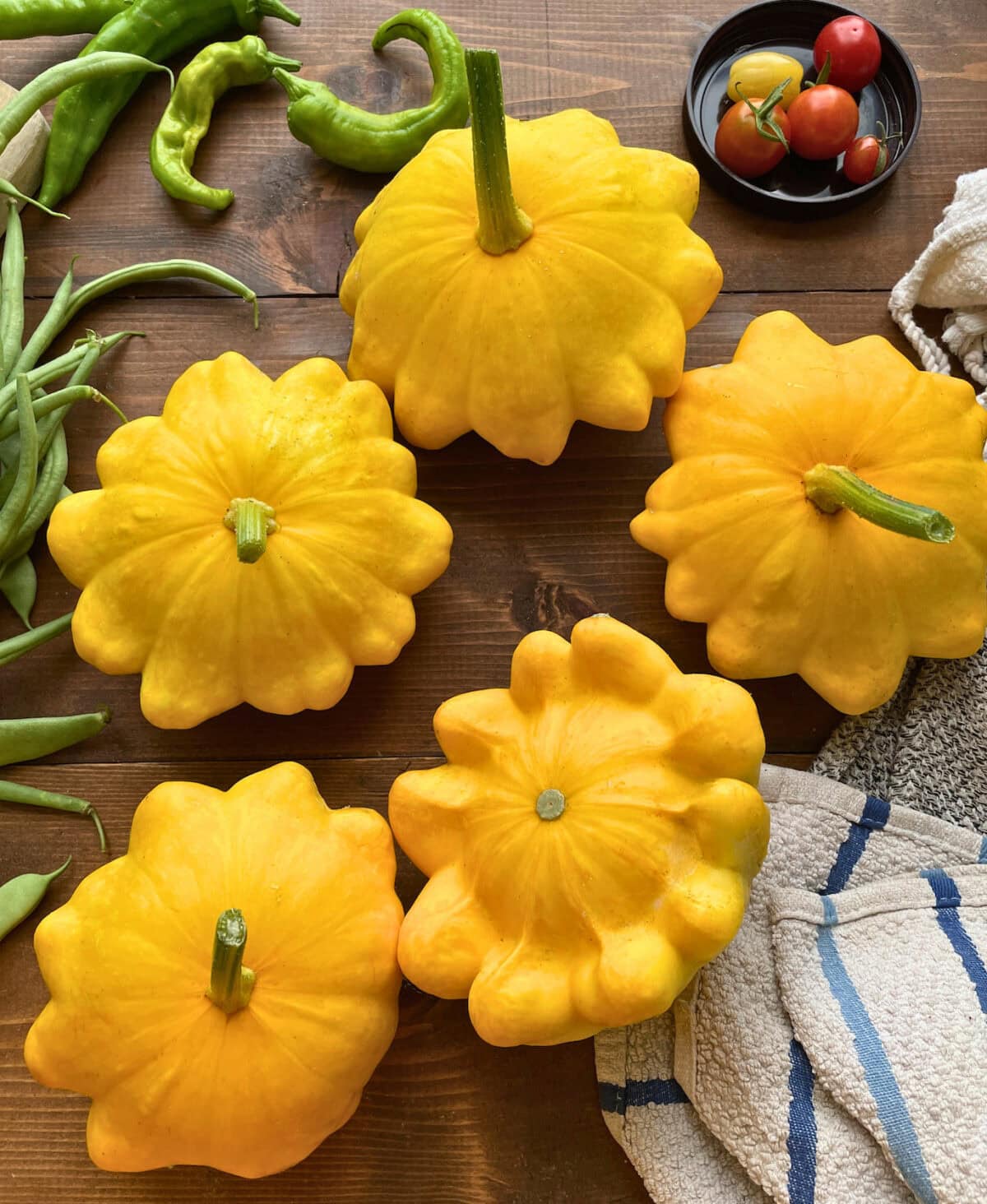 A vibrant display of yellow pattypan squash surrounded by green beans, chili peppers, and cherry tomatoes on a rustic wooden table.