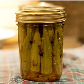 A glass jar filled with pickled okra, showcasing the vibrant green vegetables submerged in brine, resting on a checkered cloth background.