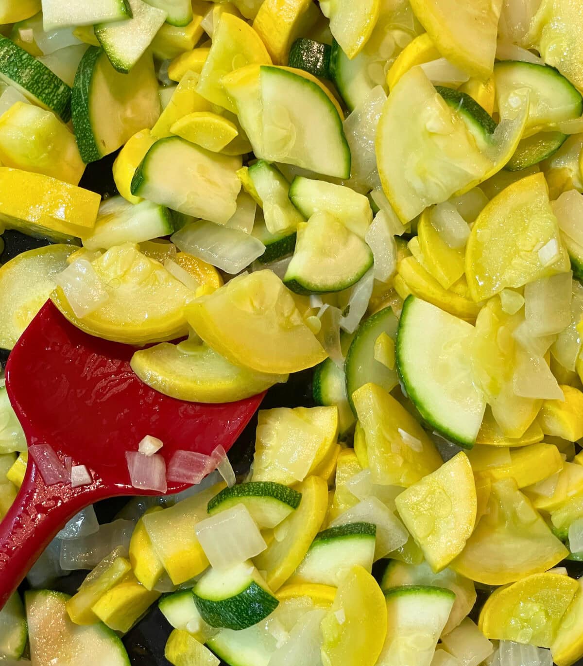 Close-up of chopped yellow and green squash mixed with onions, featuring a red spatula among the vegetables.