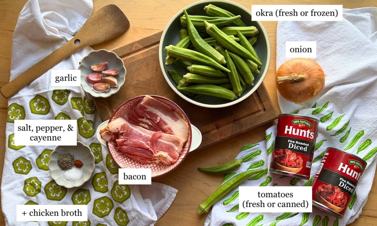 A wooden spoon, garlic, bacon, spices, chicken broth, and fresh or canned tomatoes arranged on a countertop with okra and onion.