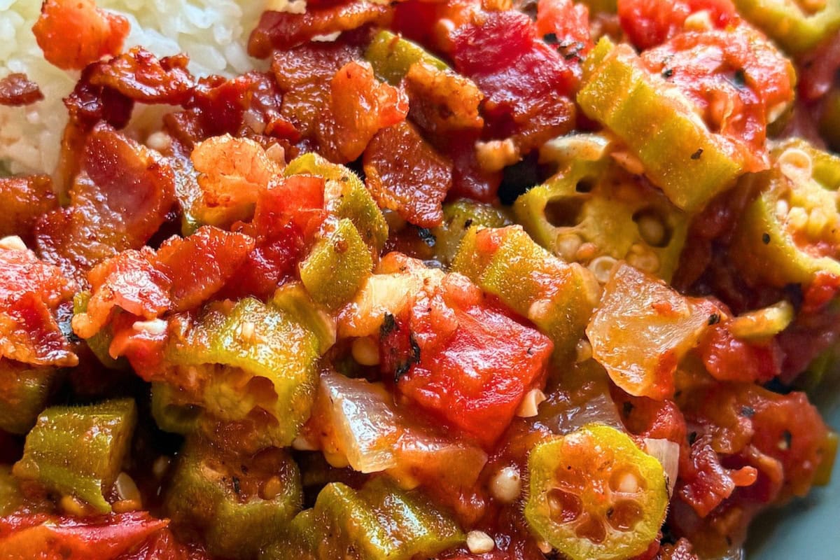 A close-up of a flavorful Southern-style dish featuring sautéed okra, tomatoes, and crispy bacon, served alongside a portion of rice.