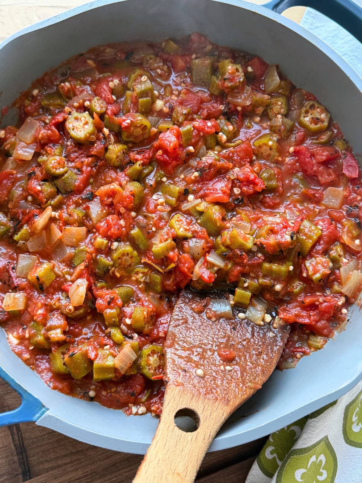 A close-up of a bubbling pot of okra and stewed tomatoes, featuring chopped okra, tomatoes, and diced onion, stirred with a wooden spoon.
