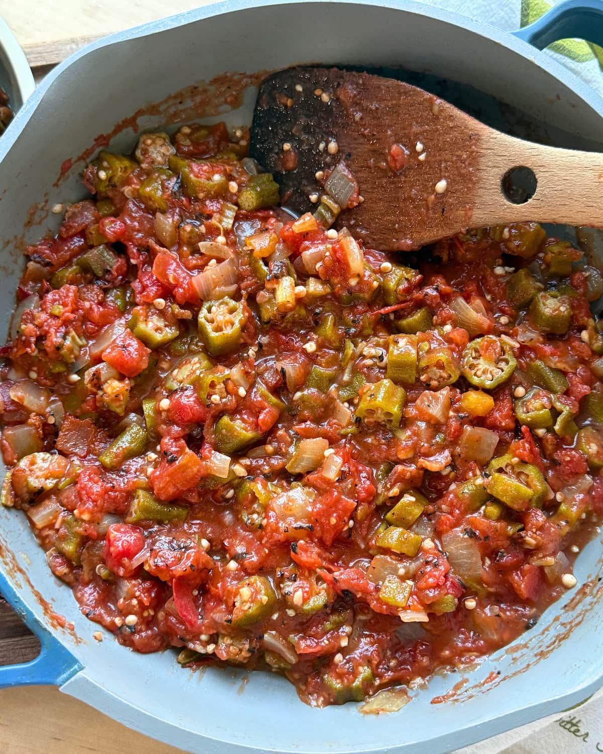 A close-up of a pot containing a colorful mixture of diced okra, tomatoes, onions, and spices, with a wooden spoon resting on the side.
