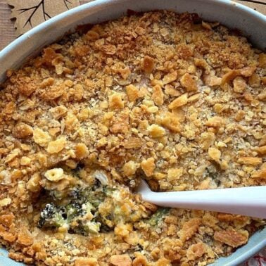 A close-up view of a baked broccoli casserole with a crunchy cornflake topping in a blue dish, featuring a serving spoon and a rustic wooden background.