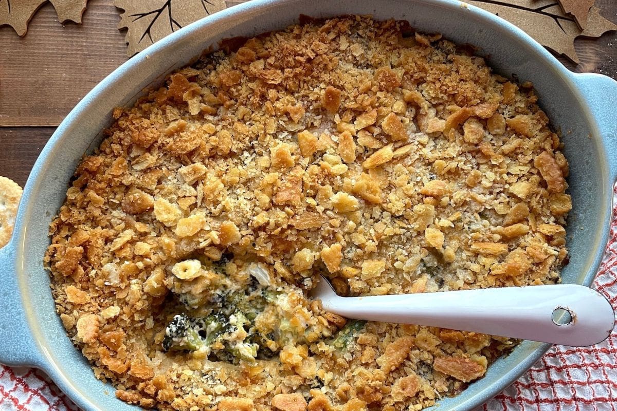 A close-up view of a baked broccoli casserole with a crunchy cornflake topping in a blue dish, featuring a serving spoon and a rustic wooden background.