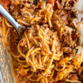 A close-up of a hearty beef spaghetti bake with melted cheese, herbs, and a fork ready to serve.