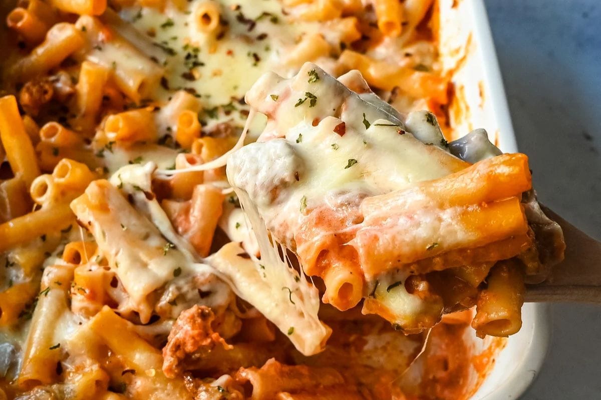 A close-up of a cheesy pasta bake with rigatoni and minced meat, being served with a wooden spatula.