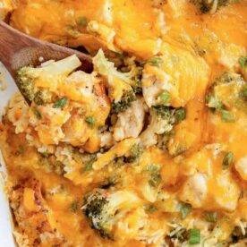 A close-up of a cheesy chicken and broccoli casserole with rice, fresh green onions, and a wooden spoon serving from a white dish.
