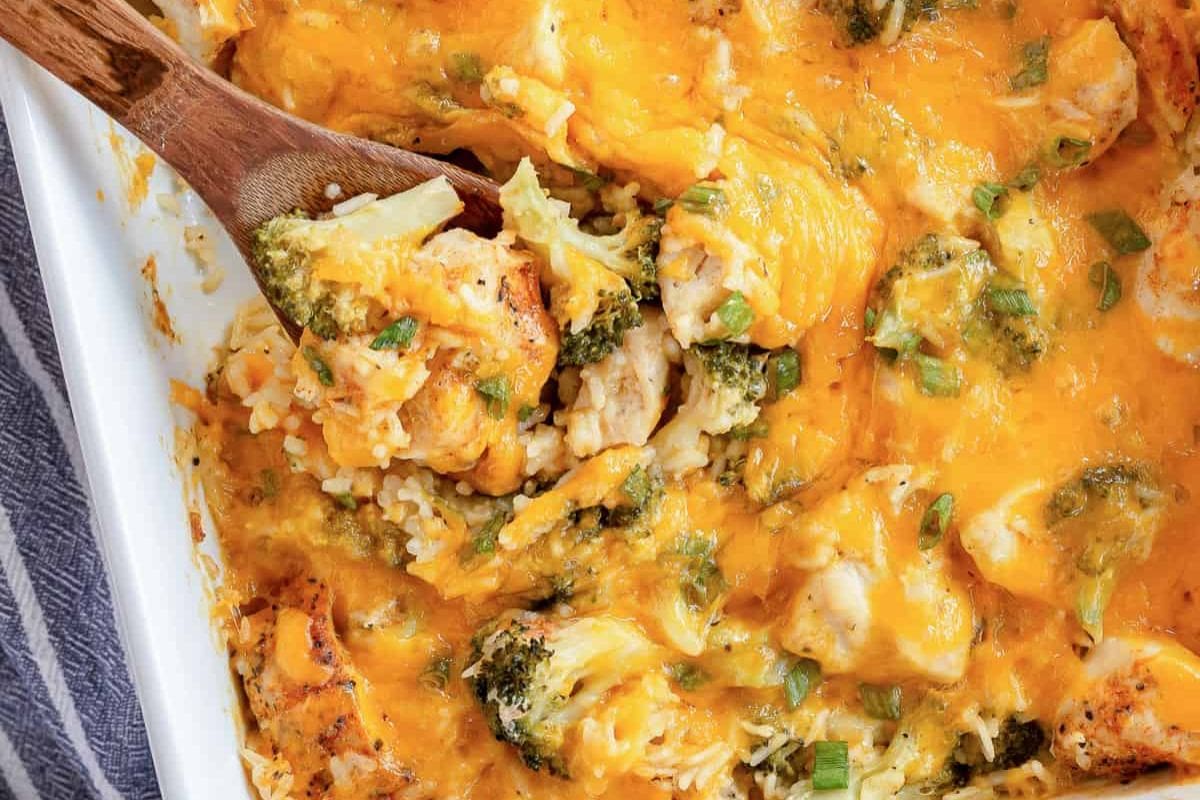 A close-up of a cheesy chicken and broccoli casserole with rice, fresh green onions, and a wooden spoon serving from a white dish.