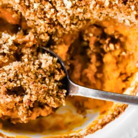 A close-up view of a spoon digging into a creamy pumpkin casserole with a crunchy streusel topping in a white baking dish.
