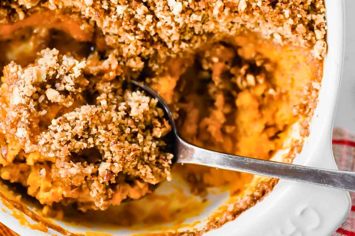 A close-up view of a spoon digging into a creamy pumpkin casserole with a crunchy streusel topping in a white baking dish.